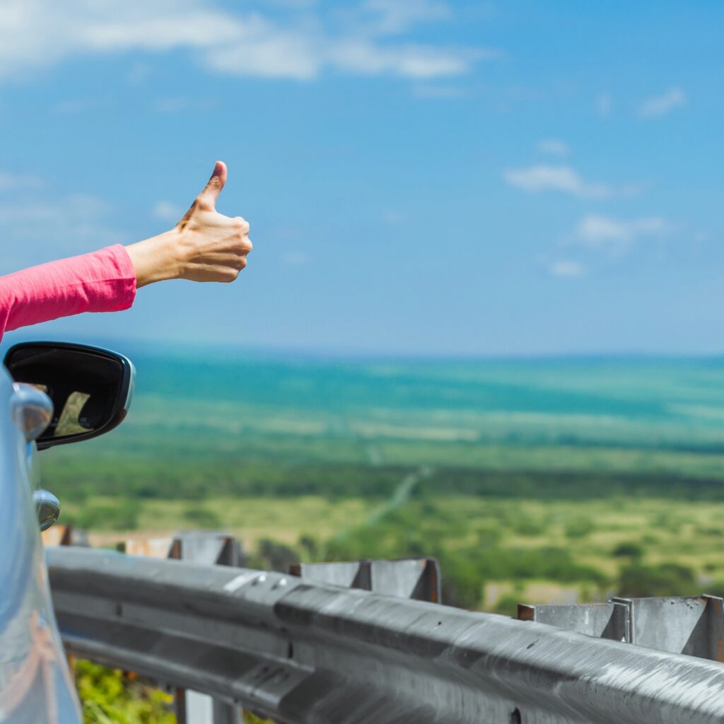 A hand giving a thumbs-up out of a car window with scenic green landscape in the background, perfect for exploring Hawaii and Hawaii travel on a budget