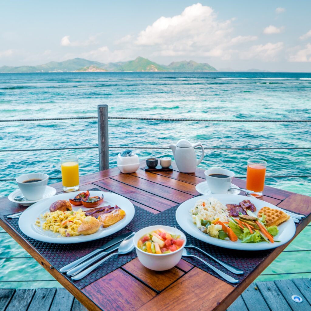 A breakfast table with plates of food and drinks overlooking the ocean, ideal for exploring Hawaii and Hawaii travel on a budget