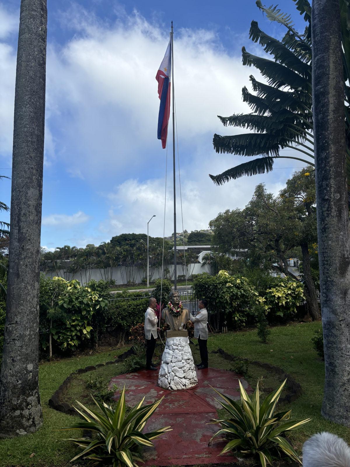 A statue of Jose Rizal with two people placing leis around it, beneath the Philippine flag, honoring Rizal's memory